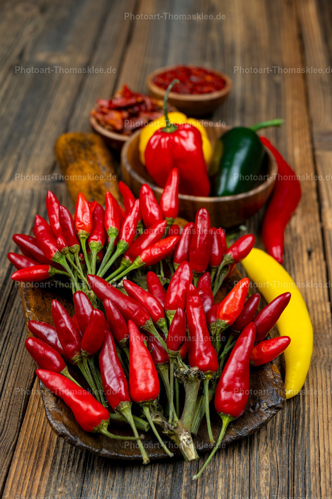 Chili-3 | Auf einem rustikalen Holztisch arrangieren sich leuchtend rote Chilischoten in einer dunklen Schale. Daneben liegen gelbe und grüne Paprika sowie weitere Gewürze in kleinen Holzschalen. Die scharfen Chilis dominieren die Komposition mit ihrer intensiven roten Farbe. Das warme Holz bildet einen natürlichen Rahmen für diese appetitliche Gewürzsammlung. Getrocknete Chilis und frische Paprika ergänzen das harmonische Arrangement perfekt. - Realisiert mit Pictrs.com