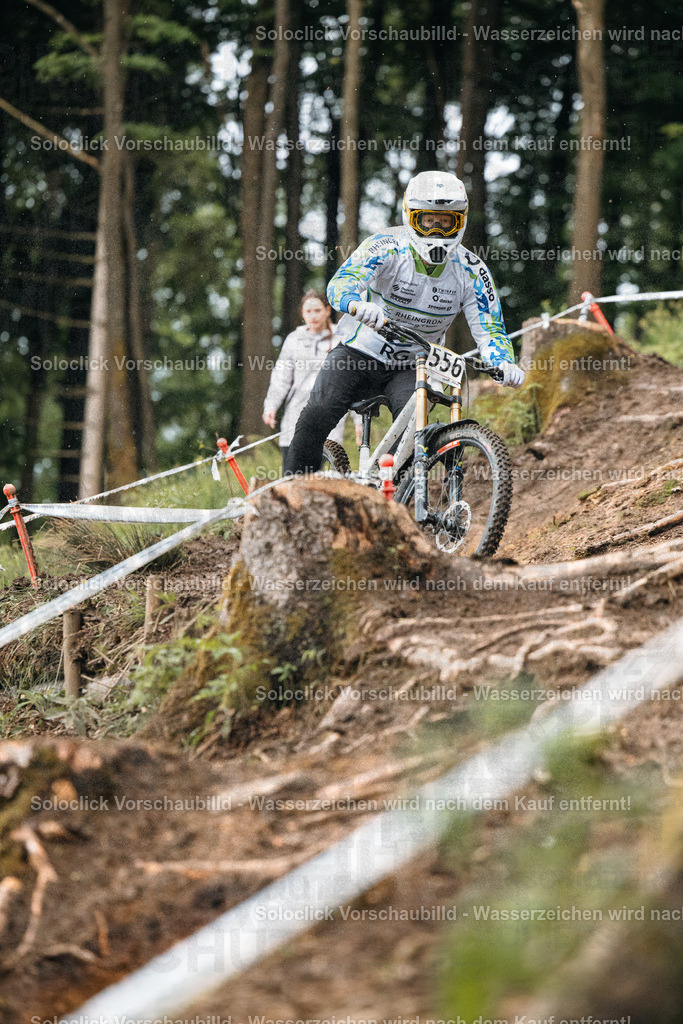Mountainbiker im Sturmlauf | Das Foto zeigt einen Mountainbiker in voller Montur, der einen steilen, bewaldeten Hang hinunterfährt. Er trägt einen Helm mit Visier, eine Weste mit Logos und Schutzkleidung. Der Hintergrund besteht aus dichtem Grün und einem Band, das den Kurs markiert. Das Bild vermittelt ein Gefühl von Geschwindigkeit und Abenteuer. - Realisiert mit Pictrs.com