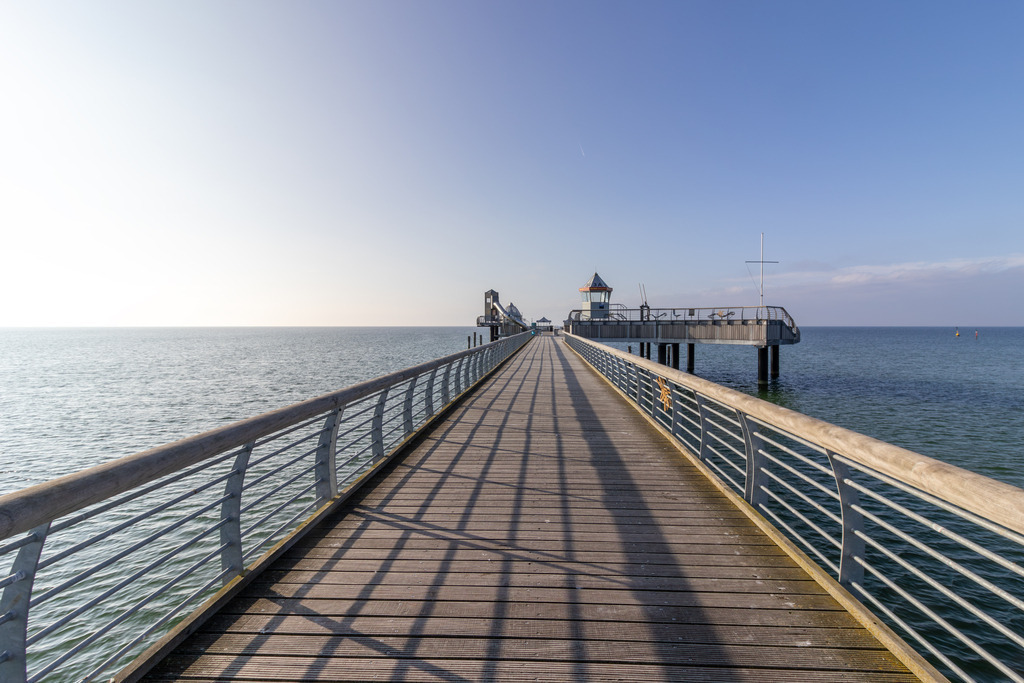 Wandbild: Seebrücke im ersten Licht – Grömitz am Morgen | Dieses Wandbild zeigt die Seebrücke von Grömitz in den frühen Morgenstunden. Ein langer Holzsteg mit seitlichen Metallgeländern führt geradlinig über den ruhigen Sandstrand hinaus auf das stille Meer. Am Ende der Brücke befindet sich ein kleines Bauwerk mit spitzem Dach – ein architektonisches Detail, das dem Bild Tiefe und Richtung verleiht. Der Himmel ist klar und zeigt einen sanften Farbverlauf von hellem zu tieferem Blau – typisch für die ersten Sonnenstrahlen des Tages. Die Komposition wirkt symmetrisch und weitläufig, ein Moment der Stille, bevor das Leben am Strand erwacht. Dieses Motiv eignet sich ideal als Wandbild für maritime Wohnkonzepte – ob als Leinwandbild, Acrylglasbild, Alu-Dibond FineArt Print oder als Akustikbild. Ein stilvoller Akzent für Wohnzimmer, Büro oder Ferienwohnung. - Realisiert mit Pictrs.com