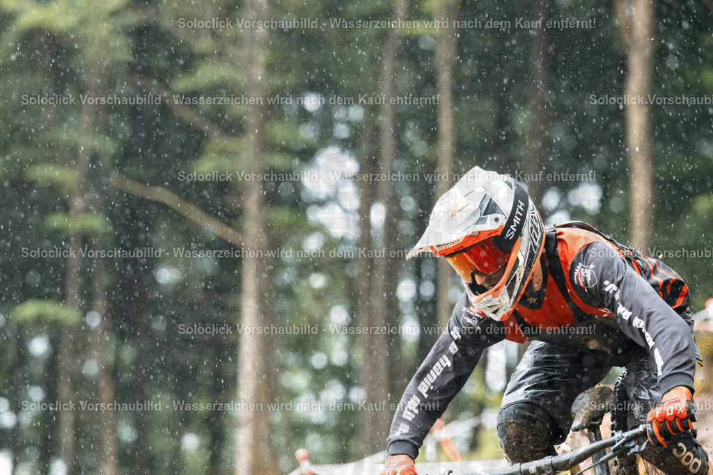 Abenteuer im Schnee | Ein Mountainbiker in voller Ausrüstung rast in einem verschneiten Wald hinunter. Er trägt einen orangefarbenen Helm mit der Aufschrift 'SMITH' und eine passende Schutzkleidung. Das Gelände ist steil und mit Schnee bedeckt, was die Fahrt anspruchsvoll macht. Der Hintergrund besteht aus dichten Bäumen, die den Eindruck eines abgelegenen und wilden Ortes verstärken. - Realisiert mit Pictrs.com