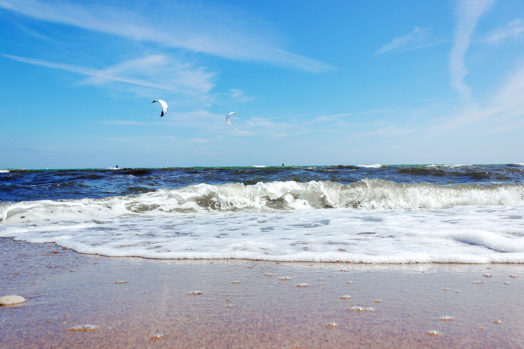 Wandbild: Strand in Weidefeld | Erleben Sie die faszinierende Küstenschönheit mit diesem Wandbild im Querformat. Es zeigt eine sich brechende Welle am sommerlichen Strand, deren Bewegung und Energie lebhaft dargestellt werden. In der Ferne sind zwei Kitesurfer zu sehen, die dem Bild eine dynamische Dimension verleihen. Der fast wolkenlose, blaue Himmel vervollständigt die Szenerie und schafft eine entspannende, maritime Atmosphäre. - Realisiert mit Pictrs.com