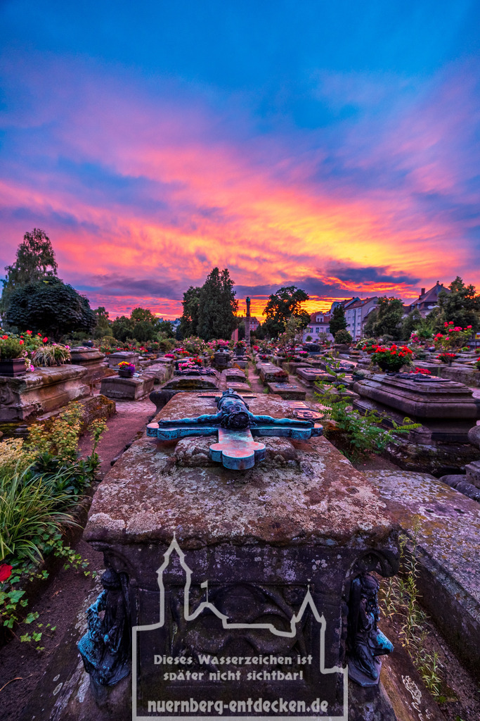 Nürnberger Stadtbild | Der Johannisfriedhof in Nürnberg bei Sonnenuntergang. Im Vordergrund steht ein altes Grab mit einem verwitterten Grabkreuz, das den gekreuzigten Jesus zeigt. Der Himmel ist in kräftigen Orange- und Violetttönen gefärbt. Die Gräber sind mit Blumen geschmückt. - Realisiert mit Pictrs.com