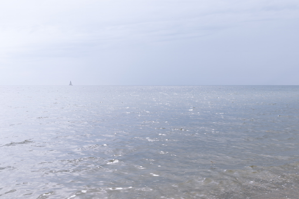 Licht über dem Meer – Minimalistisches FineArt‑Motiv mit Segelboot | Dieses FineArt‑Wandbild zeigt eine ruhige Meeresfläche unter einem hellen, leicht bewölkten Himmel. Die sanften Wellen reflektieren das Licht und verleihen der Szene eine klare, luftige Atmosphäre. In der Ferne setzt ein einzelnes Segelboot einen dezenten, eleganten Akzent, der die Weite des Meeres betont, ohne die minimalistische Bildsprache zu durchbrechen.Das Motiv wirkt zeitlos, reduziert und harmonisch – ideal für moderne, puristische Räume, die Ruhe und Offenheit ausstrahlen sollen.