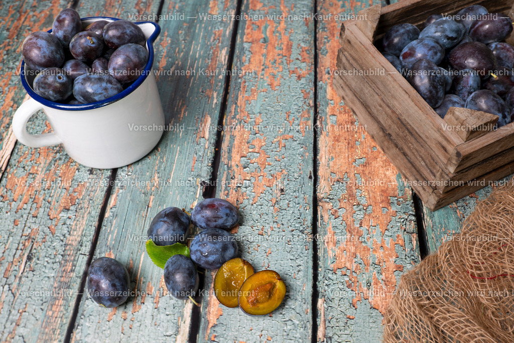 Eine Emailletkanne und eine Holzkiste voll mit erntefrischenn Pflaumen | Eine Emailletkanne und eine Holzkiste voll mit erntefrischenn Pflaumen auf alter, rustikaler Holztischplatte. Eine Pflaume aufgeschnitten. - Realisiert mit Pictrs.com