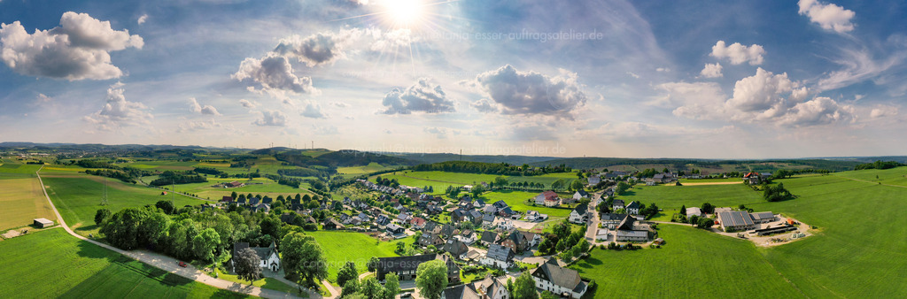 Panoramafoto Wülfte bei Brilon von oben | Luftbild Panorama des Ortsteil Wülfte bei Brilon im Sauerland. Häuser, Felder, Bauernhöfe und eine Kapelle vor grünen Hügeln. Blauer Himmel mit Sonnenschein.