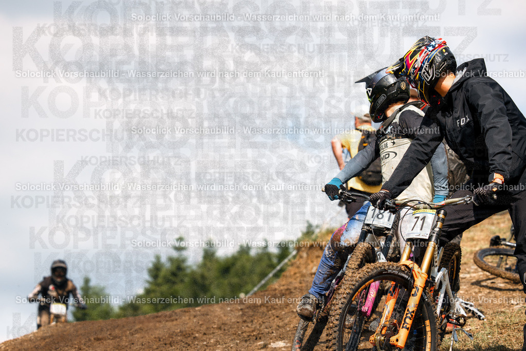 Bereit für den Abstieg | Das Bild zeigt drei Mountainbiker in voller Ausrüstung, die sich auf einem staubigen Hügel positionieren, bereit für den Start eines Downhill-Rennens. Die beiden vorderen Fahrer stehen im Fokus, während der dritte Fahrer im Hintergrund zu sehen ist. Die Landschaft ist geprägt von einem staubigen Hügel mit vereinzelten Büschen und einem Blick auf das Meer im Hintergrund. - Realisiert mit Pictrs.com