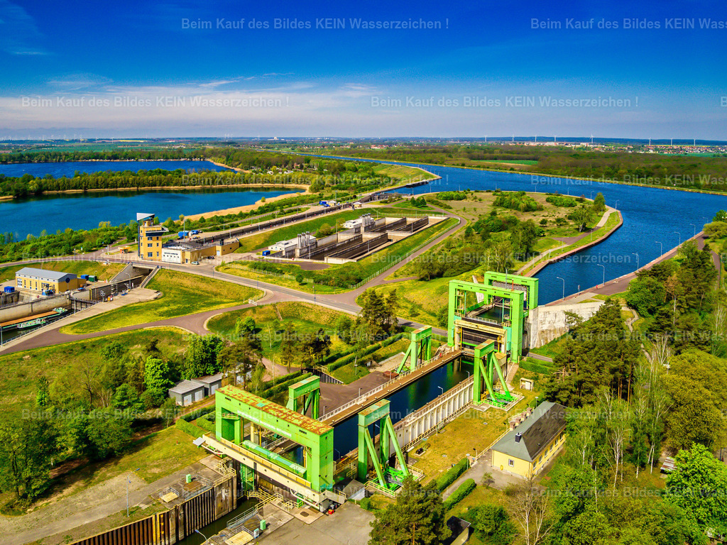Schiffshebewerk Rothensee Doppelsparschleuse Barleber See-2 | Diese farbenfrohe Luftaufnahme zeigt das historische Schiffshebewerk Rothensee an einem sonnigen Sommertag. Die markante Stahlkonstruktion steht im Mittelpunkt des Bildes und dokumentiert eines der bedeutendsten technischen Denkmale Deutschlands. Im Hintergrund ist die moderne Doppelsparschleuse Rothensee zu erkennen – ein eindrucksvoller Kontrast zwischen historischer Ingenieurbaukunst und moderner Wasserstraßentechnik. Aus der Luft werden die Dimensionen der gesamten Anlage und ihre Einbindung in das Wasserstraßenkreuz Magdeburg besonders deutlich.Das Schiffshebewerk wurde 1938 in Betrieb genommen und galt damals als technische Meisterleistung. Sein 85 Meter langer und 12 Meter breiter Trog hebt Schiffe – abhängig vom Wasserstand der Elbe – um bis zu **18,46 Meter** an. Das bewegte Gesamtgewicht des mit Wasser gefüllten Troges beträgt rund **5.400 Tonnen**. Besonders bemerkenswert ist das Zwei-Schwimmer-System: Zwei gewaltige, luftgefüllte Schwimmkörper mit einer Höhe von 36 Metern tragen den Trog nahezu im Gleichgewicht, sodass für den Hubvorgang vergleichsweise wenig Energie benötigt wird.Heute ist das Schiffshebewerk nicht nur ein einzigartiges Denkmal der Industriekultur, sondern auch das **letzte betriebsfähige Zwei-Schwimmer-Schiffshebewerk Europas**. Gemeinsam mit der benachbarten Doppelsparschleuse und der Trogbrücke bildet es das Herzstück des Wasserstraßenkreuzes Magdeburg und zählt zu den beeindruckendsten Ingenieurbauwerken der europäischen Binnenschifffahrt. - Realisiert mit Pictrs.com