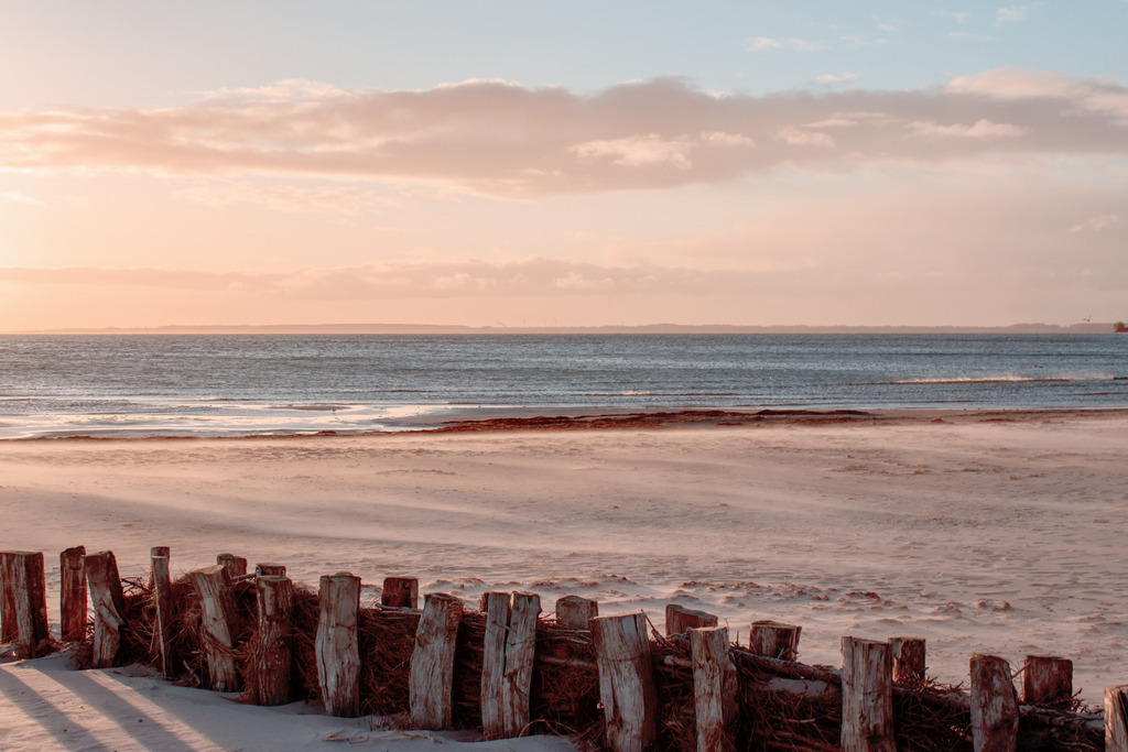 Wandbild: Morgenrot am Meer in dezenten Farben | Dieses Wandbild im Querformat zeigt eine wunderschöne Morgenstimmung am Meer. Das ganze Bild ist durch den Sonnenaufgang leicht rötlich. Im Vordergrund steht ein Sandfang. Auf der linken Seite scheint die Sonne durch einige Pfähle und erzeugt dort Schatten. Der Himmel ist blau und rotorange. Die Farben wirken alle sehr dezent. Holen Sie sich diese schöne maritime Morgenstimmung auf Leinwand, Aluminium-Platte oder als Glasbild. Ideal fürs Wohnzimmer, Schlafzimmer, Küche, den Arbeitsplatz oder die Ferienwohnung. Die Wandbilder werden individuell für Sie in vielen Abmessungen produziert. - Realisiert mit Pictrs.com