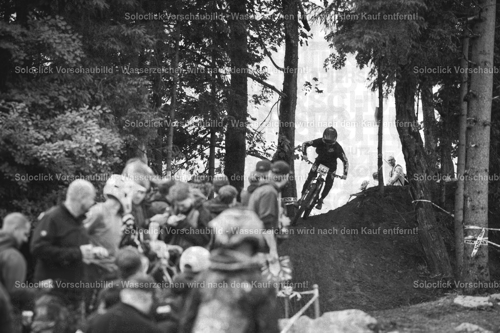 Mountainbike-Action im Wald | Das Schwarz-Weiß-Foto zeigt einen Mountainbiker in voller Montur, der in der Luft über eine Erhebung springt. Er trägt einen Helm, eine Brille und Schutzkleidung. Im Hintergrund ist ein Publikum zu sehen, das den Sprung verfolgt. Die Szene spielt sich in einem bewaldeten Gebiet statt, möglicherweise bei einem Mountainbike-Wettbewerb. - Realisiert mit Pictrs.com