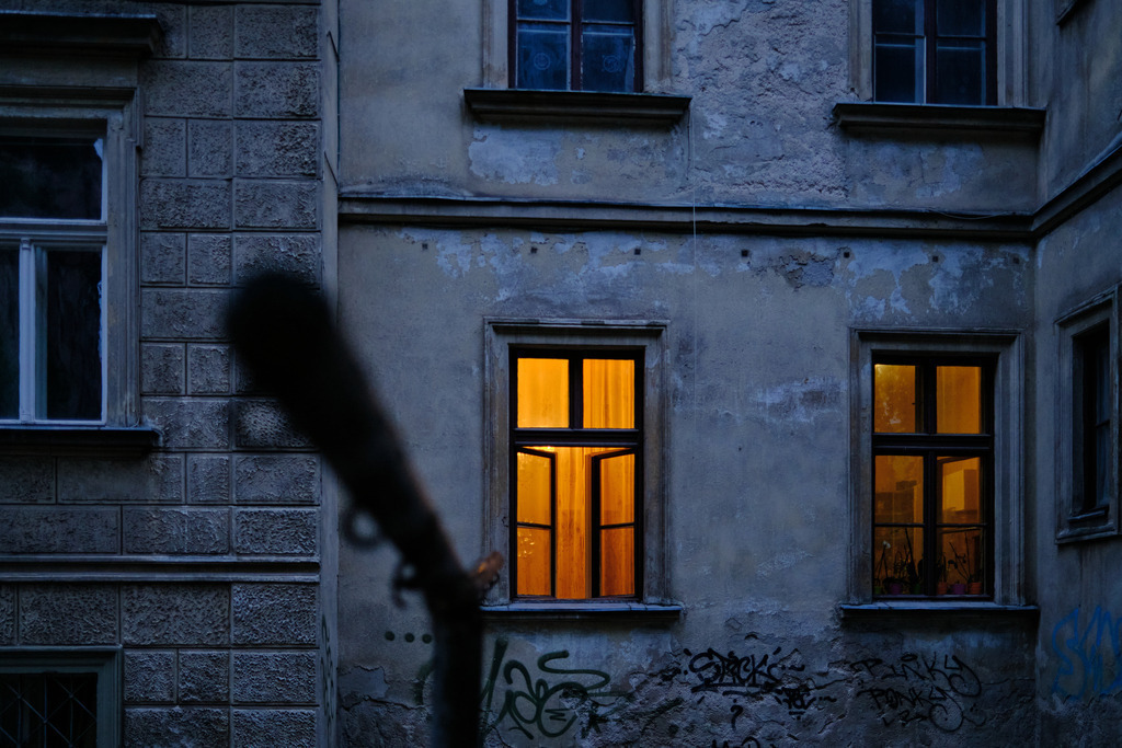 Wohnhaus mit beleuchtetem Fenster am Abend | Ljubljana, Slowenien - June 22, 2025: Wohnhaus mit beleuchtetem Fenster am Abend, im Vordergrund eine Teppichstange. - Realisiert mit Pictrs.com