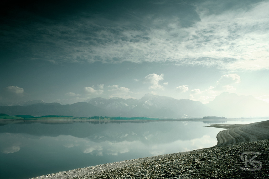 Stille Symmetrie – Perfekte Spiegelung am See unter dramatischem Himmel | Minimalistische Seelandschaft mit perfekter Wasserspiegelung unter dramatischem Wolkenhimmel. Bergsilhouette und atmosphärisches Licht schaffen eine symmetrische, meditative Komposition von Stefan Kuhn. - Realisiert mit Pictrs.com