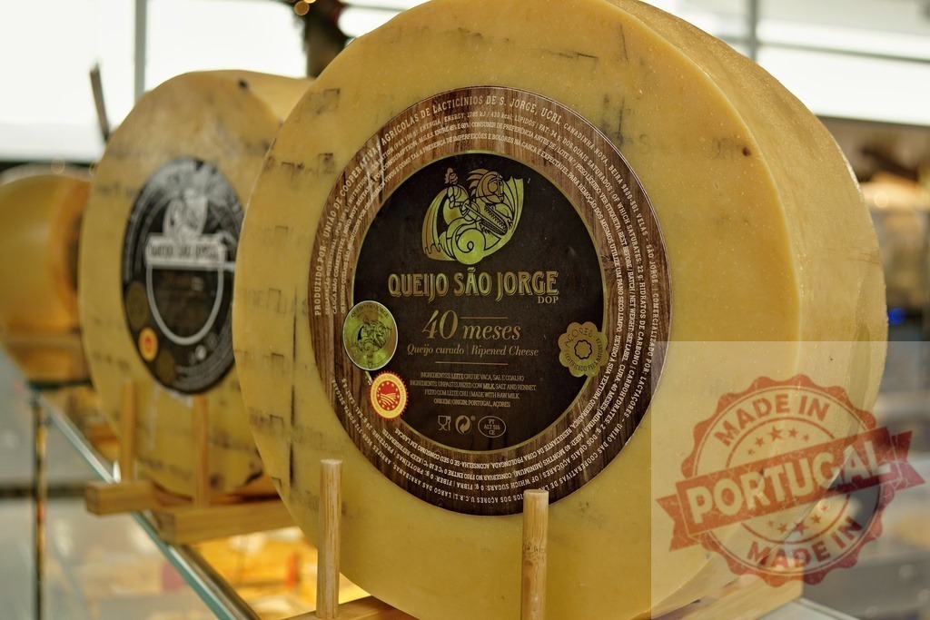 Queijo São Jorge cheese wheels displaying labels in market | Queijo São Jorge DOP cheese wheels aging, displayed in a store - Realized with Pictrs.com