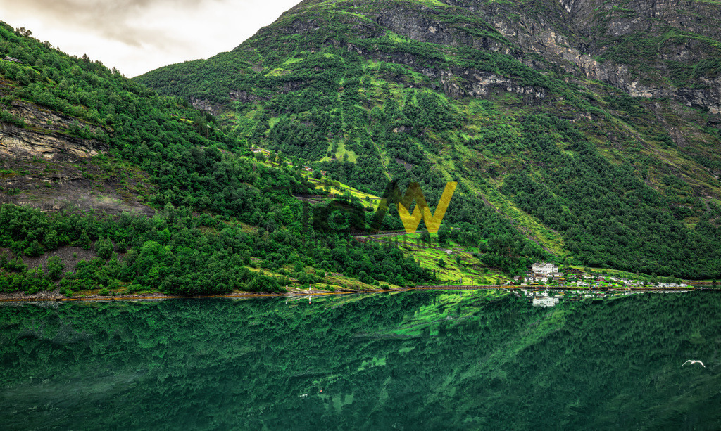 Der Ort Geiranger-im Geirangerfjord in Norwegen-Spiegelung | Der Fjord ist Teil des UNESCO-Weltnaturerbes und für seine dramatische Naturlandschaft bekannt. Er ist von bis zu 1.700 Meter hohen Bergen umgeben, die steil in das tiefblaue Wasser abfallen und einen markanten Kontrast zur grünen Vegetation bilden. Die Region zeichnet sich durch majestätische Berge, tiefe Täler und zahlreiche Wasserfälle aus, darunter die berühmten "Sieben Schwestern". Am Ende des etwa 15 km langen Fjords liegt das malerische Dorf Geiranger, ein beliebter Ausgangspunkt für Wanderungen und Erkundungen.  - Realisiert mit Pictrs.com