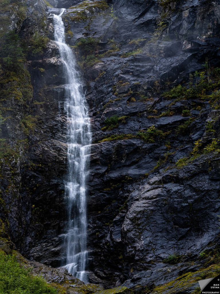 Waterfall on the Shores of Fjærlandsfjord | During my walk along the western shore of Fjærlandsfjord, I came across several waterfalls of different sizes. This one caught my attention from a distance, cascading down a steep wall of dark, rain-soaked rock. The smooth, polished surfaces surrounding the falls suggested that there must be times when far more water rushes down the mountainside, turning this relatively slender cascade into a much broader and more powerful waterfall. - Realisiert mit Pictrs.com