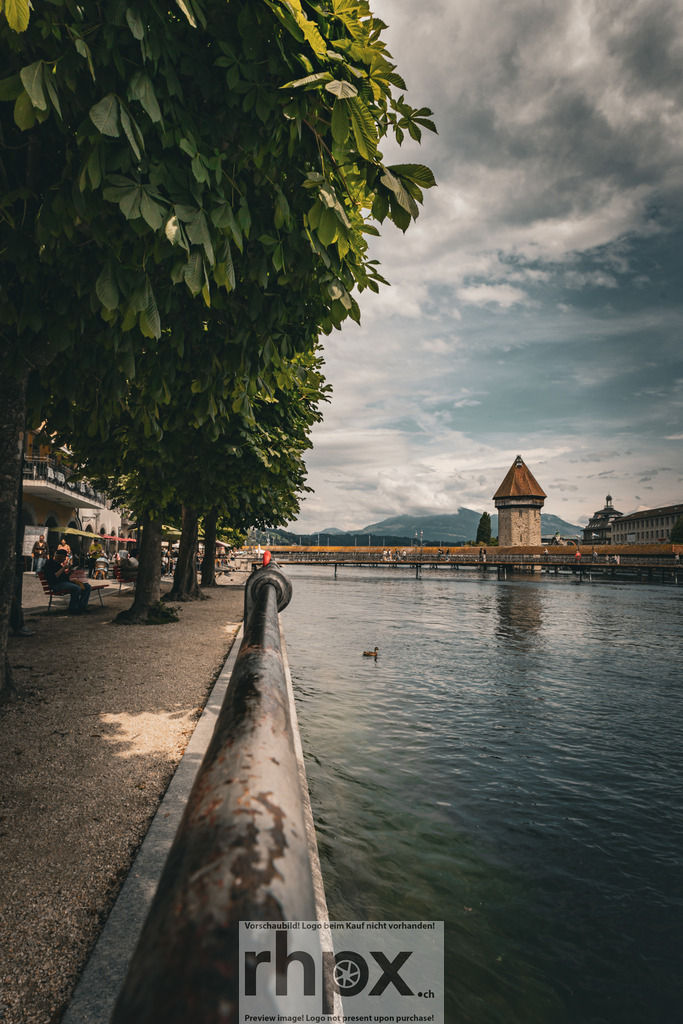Sommer am Reussufer | Schatten. Fluss. Wahrzeichen.Ein entspannter Spaziergang unter den schattigen Kastanienbäumen entlang der Reuss eröffnet diesen klassischen Blick auf die Kapellbrücke. Die friedliche Stimmung am Wasser und das ferne Bergpanorama fangen das sommerliche Lebensgefühl in Luzern perfekt ein.Wähle unter "Produktauswahl" dein Wunschformat: Vom klassischen Wandbild über Puzzle &amp; Tassen bis zum digitalen Download (z.B. als Handy-Hintergrund). - Realisiert mit Pictrs.com