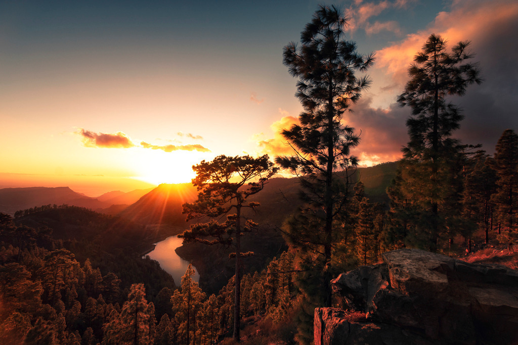 Sonnenuntergang über den Bergen – Gran Canaria | Ein goldener Sonnenuntergang taucht die Landschaft von Gran Canaria in warmes Licht. Die typischen kanarischen Kiefern zeichnen sich als Silhouetten vor dem leuchtenden Himmel ab, während sich die letzten Sonnenstrahlen über die Berge und das Tal mit dem geschwungenen Stausee legen. Ein Moment der Stille, der die wilde Schönheit dieser Insel eindrucksvoll zur Geltung bringt. - Realisiert mit Pictrs.com