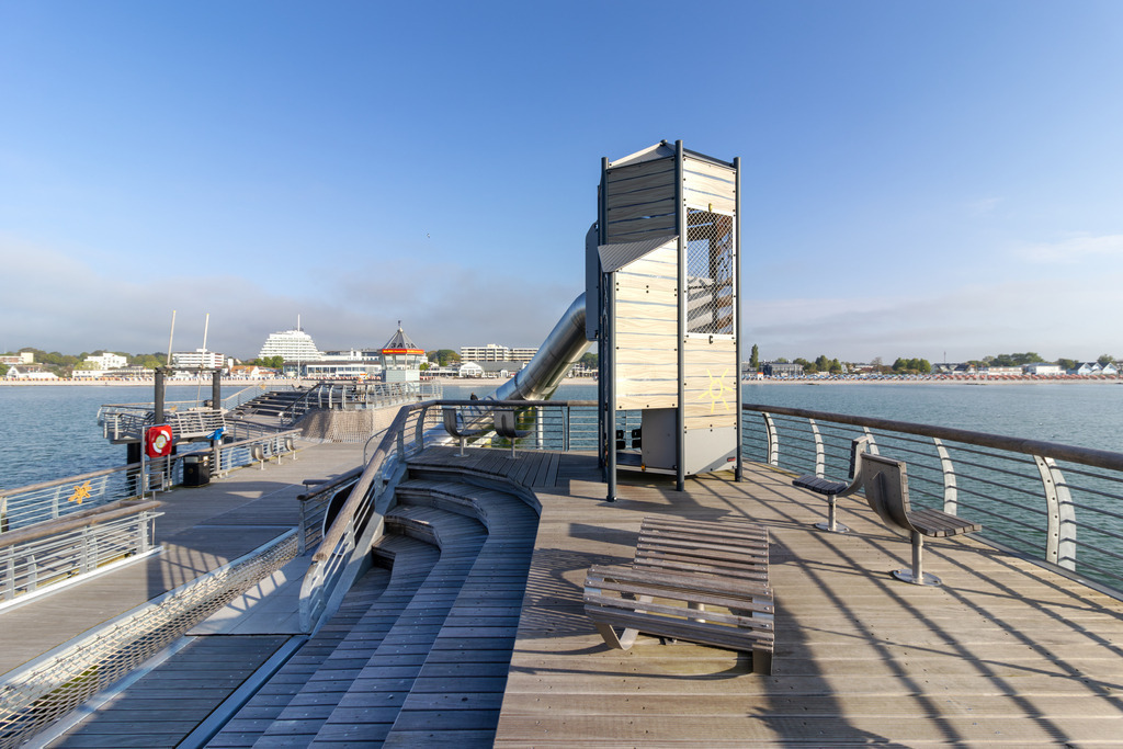 Wandbild: Spiel und Meer – Kletterturm auf der Seebrücke Grömitz | Dieses Wandbild zeigt eine besondere Perspektive von der Seebrücke in Grömitz: Im Mittelpunkt steht ein moderner Kletterturm mit integrierter Tunnelrutsche – ein Highlight für Familien und Kinder direkt über dem Wasser. Die Konstruktion aus Holz und Metall fügt sich harmonisch in die Umgebung ein und verbindet Freizeitspaß mit maritimer Architektur. Entlang der Seebrücke laden Sitzbänke zum Verweilen ein, während das ruhige Meer und der klare Himmel eine entspannte Kulisse schaffen. Im Hintergrund sind Strand und Gebäude zu erkennen, die das Bild als typisches Küstenmotiv abrunden. Dieses Motiv eignet sich ideal als Wandbild für maritime Wohnkonzepte oder als dekorativer Akzent in Ferienwohnungen, Kinderzimmern oder Aufenthaltsbereichen. Erhältlich als Leinwandbild, Acrylglasbild, Alu-Dibond FineArt Print oder als Akustikbild. - Realisiert mit Pictrs.com