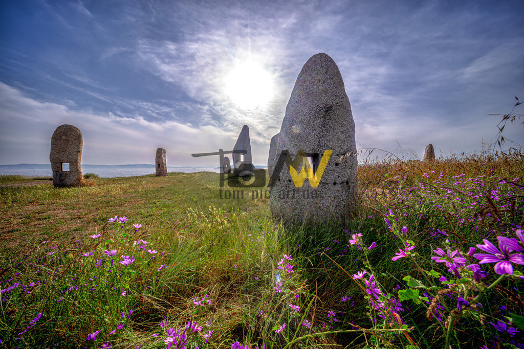 Die Menhires por la Paz in A Coruña, Galizien, Spanien | Die abgebildeten Steinskulpturen sind die "Menhire für den Frieden" (Menhires por la Paz) in A Coruña, Galizien, Spanien. Die Gruppe besteht aus zwölf Skulpturen, die 2003 vom galizischen Bildhauer Manolo Paz geschaffen und eingeweiht wurden. Sie befinden sich im Skulpturenpark des Herkulesturms (Parque Escultórico da Torre de Hércules) auf dem Campo de la Rata. Die Skulpturen repräsentieren die Familie und haben kleine Fenster, die unterschiedliche Ausblicke auf die Umgebung, das Meer und den nahe gelegenen Herkulesturm bieten.  - Realisiert mit Pictrs.com