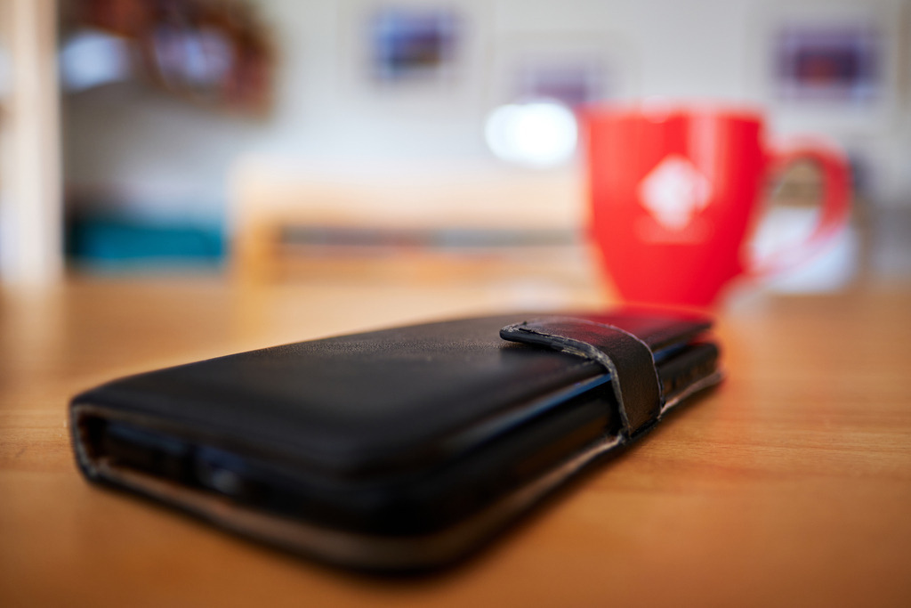 Handy mit Hülle liegt auf dem Tisch, dahinter ein roter Kaffeebecher | Wien, Austria - March 17, 2021: Handy mit Hülle liegt auf dem Tisch, dahinter ein roter Kaffeebecher. - Realisiert mit Pictrs.com