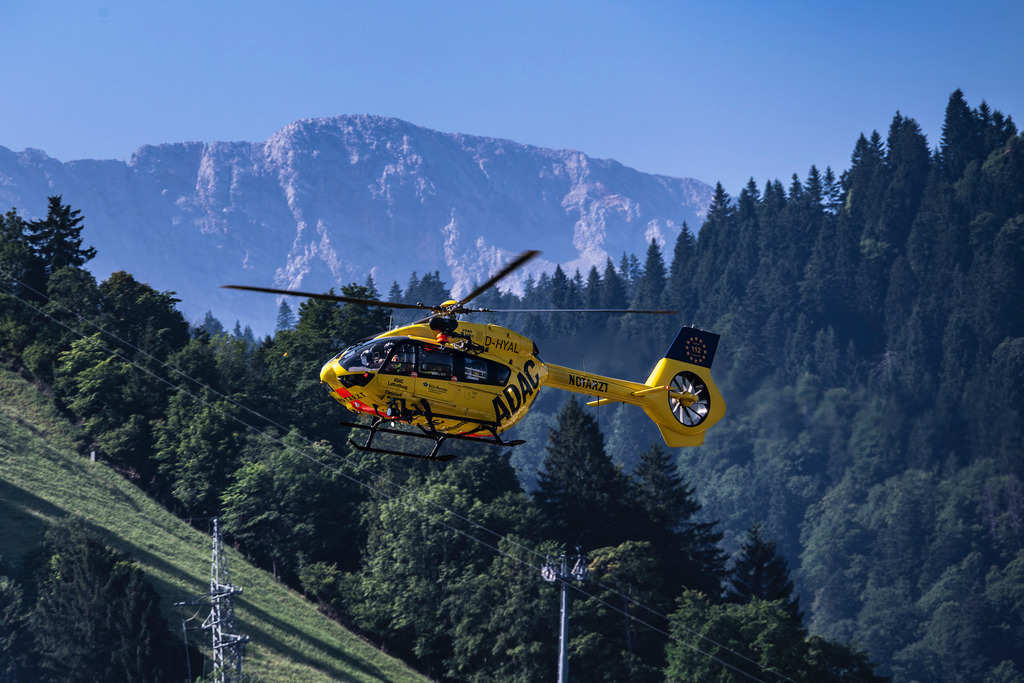 ADAC Rettungshubschrauber in den Alpen | Ein markanter gelber ADAC-Rettungshubschrauber (Christoph 1, Kennung D-HYAL) schwebt in einer dynamischen Vorwärtsbewegung vor einer imposanten Bergkulisse. Die schroffen, hellgrauen Felswände des Massivs bilden einen majestätischen Kontrast zum tiefgrünen, dicht bewaldeten Hang im Vordergrund, während die klare Bergluft die Szene in ein kühles, natürliches Licht taucht. - Realisiert mit Pictrs.com