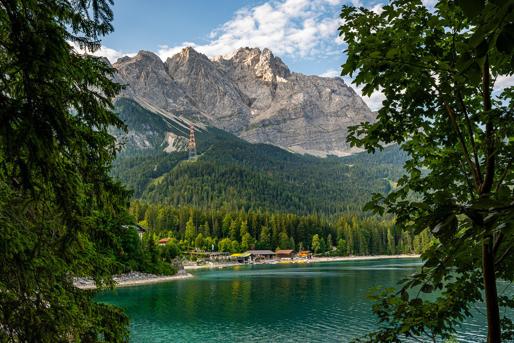 Alpenidylle pur: Der Eibsee unter der Zugspitze | Eingerahmt von dunklen Nadelbäumen eröffnet sich ein atemberaubender Blick auf den smaragdgrünen Eibsee und das mächtige Massiv der Zugspitze. Das Foto fängt die tiefe Ruhe und erhabene Schönheit der Natur ein. Man riecht förmlich die frische Bergluft und spürt die kühle Brise, die über das kristallklare Wasser streicht. Ein kleiner Sendemast auf halber Höhe erinnert an die Zivilisation, während die markanten Felswände der Zugspitze den Himmel berühren. Ein perfekter Ort, um die Seele baumeln zu lassen. - Realisiert mit Pictrs.com