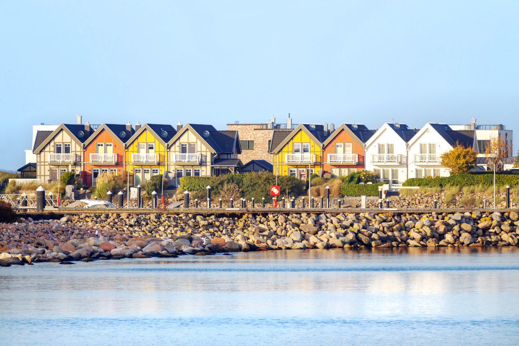 Leinwandbild Hafenglück – Bunte Giebelhäuser im Ostseeresort Olpenitz | Skandinavisches Flair an der Schlei-Mündung: Dieses Wandbild zeigt die markanten, farbenfrohen Ferienhäuser im OstseeResort Olpenitz. Die Spiegelung der bunten Fassaden im ruhigen Wasser und die massive Steinmole fangen die moderne maritime Architektur dieses einzigartigen Resorts perfekt ein. Das klare Licht und die harmonische Farbstimmung bringen ein Gefühl von Lifestyle und nordischer Lebensfreude direkt in Ihre Räume. Hochwertig auf Leinwand gedruckt, ist dieses Motiv ein Highlight für alle Fans der Region rund um Kappeln und Liebhaber zeitgemäßer Küstenarchitektur. - Realisiert mit Pictrs.com