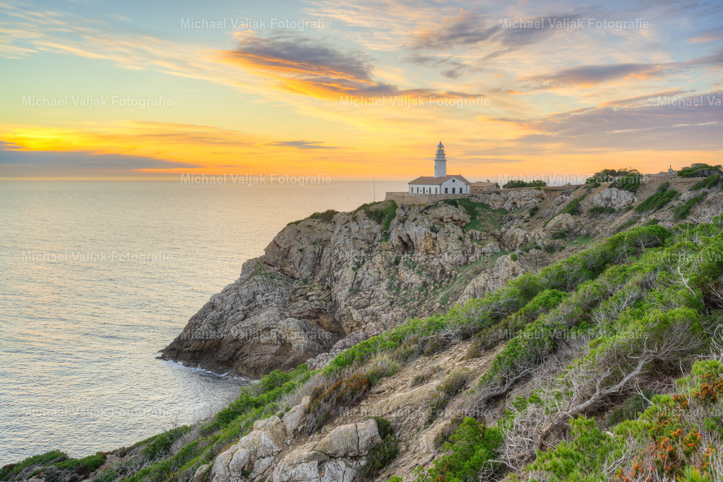 Morgenstimmung am Faro de Capdepera auf Mallorca | Der Faro de Capdepera steht zum Sonnenaufgang in einer Landschaft, die durch ihre Küstenform fast mehr erzählt als der Leuchtturm selbst. Die warmen Rot‑ und Orangetöne des Himmels legen sich über die zerklüfteten Felsen, die sich klar und strukturiert bis zum Meer hinunterziehen. Die Küste wirkt gleichzeitig rau und ruhig, vom ersten Licht weich betont, ohne an Präsenz zu verlieren. Der Leuchtturm fügt sich als heller Punkt in diese Szenerie ein, aber die eigentliche Wirkung entsteht durch die Kombination aus warmem Morgenlicht, offener See und der markanten Felslinie, die den Blick entlang der Küste führt. - Realisiert mit Pictrs.com