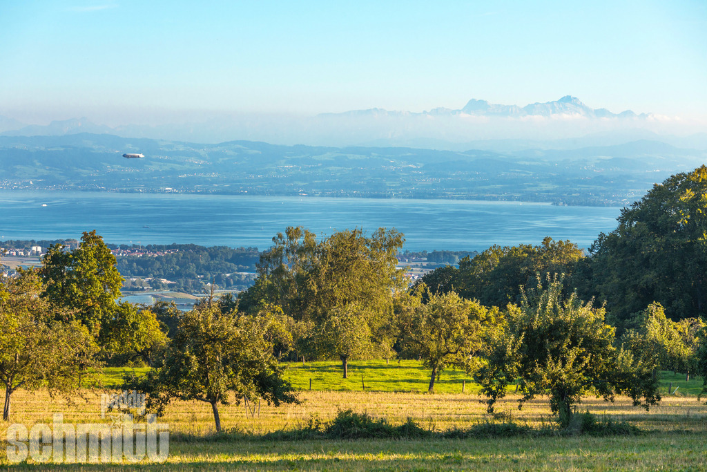 Zeppelin über dem Bodensee mit Blick auf den Säntis bei Marktdorf | Vom Gehrenberg aus eröffnet sich ein beeindruckender Blick über den Bodensee bis zum markanten Säntis. Ein Zeppelin schwebt durch den weiten Himmel und verbindet sich mit der faszinierenden Kulisse aus See, Alpen und Landschaft zu einem außergewöhnlichen Fotomotiv. - Realisiert mit Pictrs.com