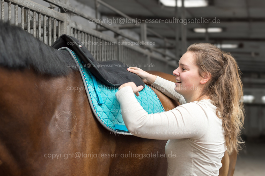 Woman saddles horse, thick padded pad | Eine junge Frau sattelt ihr Pferd auf dem Reitplatz. Sie legt das dick gepolsterte Pad an. Der Reitstall ist unscharf im Hintergrund zu sehen.