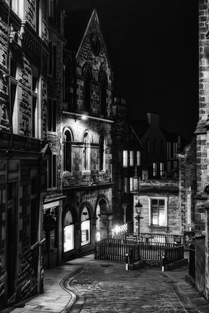 Nacht in Edinburghs Upper Bow | Eine enge, gepflasterte Gasse in Edinburghs Upper Bow schlängelt sich durch historische Steingebäude bei Nacht. Die Szene wird von Straßenlaternen und beleuchteten Fenstern erhellt, die starke Kontraste und tiefe Schatten auf die rauen Fassaden werfen. Die Schwarz-Weiß-Darstellung betont die architektonischen Details und die atmosphärische Tiefe der urbanen Landschaft. - Realisiert mit Pictrs.com