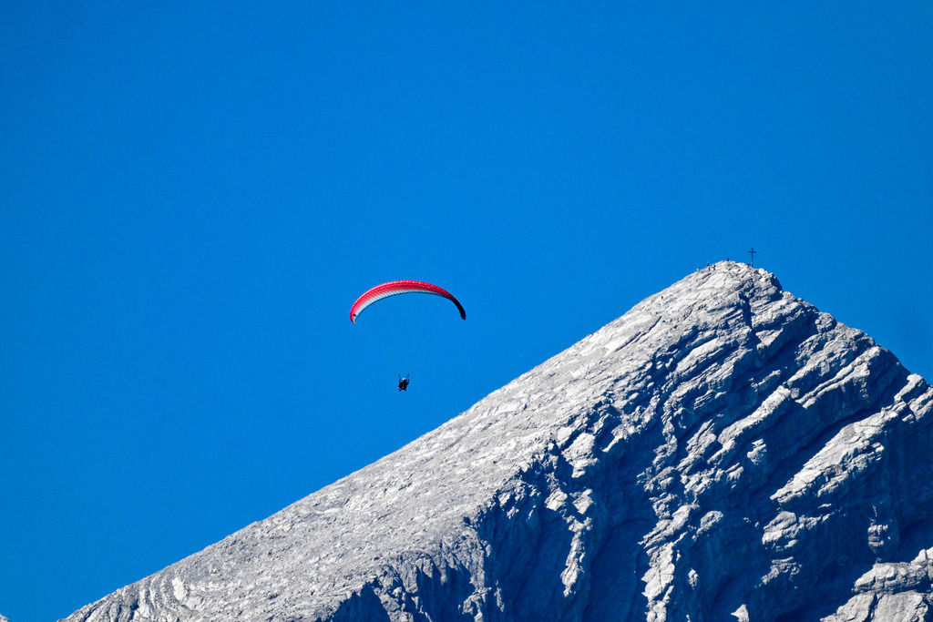 Dem Himmel so nah: Freiheit im Wind der Alpen | Ein Anblick, der den Atem raubt und das Gefühl grenzenloser Freiheit spürbar macht: Elegant gleitet ein Paraglider mit seinem leuchtend rot-weißen Schirm dicht an der gewaltigen Flanke der Alpspitze entlang. Die schroffen, von der Sonne hell erleuchteten Kalksteinfelsen des markanten Berggipfels ziehen im steilen Winkel nach oben, gekrönt vom fernen Gipfelkreuz. Vor dem makellosen, tiefen Blau des Sommerhimmels wird die Silhouette des Fliegers zum Symbol für den menschlichen Mut und die Sehnsucht, die Welt aus der Adlerperspektive zu erleben. Ein Bild voller Dynamik, erhabener Stille und alpiner Faszination. - Realisiert mit Pictrs.com