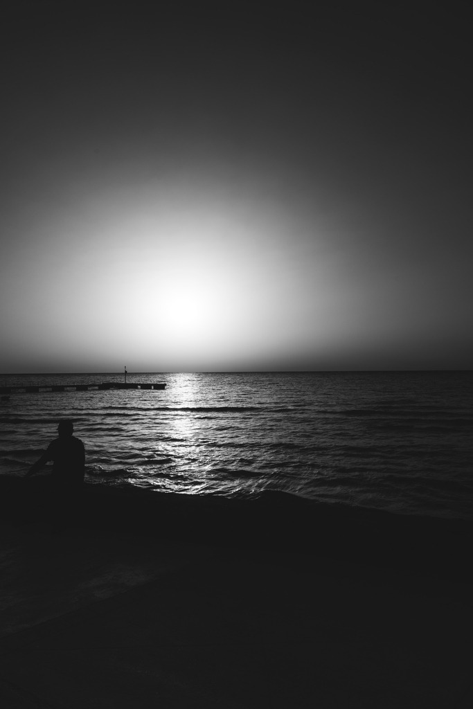 Einsamer Sonnenaufgang am Meer in Cala Millor auf Mallorca | Eine einzelne Silhouette am Ufer des Mittelmeers, während die aufgehende Sonne das Wasser vor Cala Millor zum Leuchten bringt.Die Schwarz-Weiß-Bearbeitung reduziert den Sonnenaufgang auf Licht, Reflexion, Meer und menschliche Präsenz. Der große offene Himmel und die einsame Figur verleihen der Aufnahme eine stille, fast meditative Atmosphäre.Eine einsame Silhouette am Mittelmeer während des Sonnenaufgangs in Cala Millor auf Mallorca. Licht, Wasser und weiter Himmel verbinden sich zu einem stillen Schwarz-Weiß-Motiv voller Ruhe und Atmosphäre. - Realisiert mit Pictrs.com