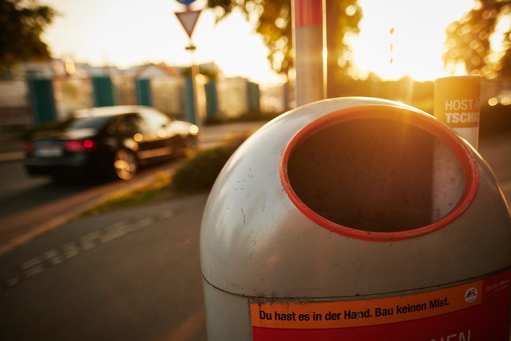 Mistkübel auf dem Gehweg | Wien, Austria - September 20, 2020: Mistkübel auf dem Gehweg im Gegenlicht, im Hintergrund ein Auto mit Strasse. - Realisiert mit Pictrs.com