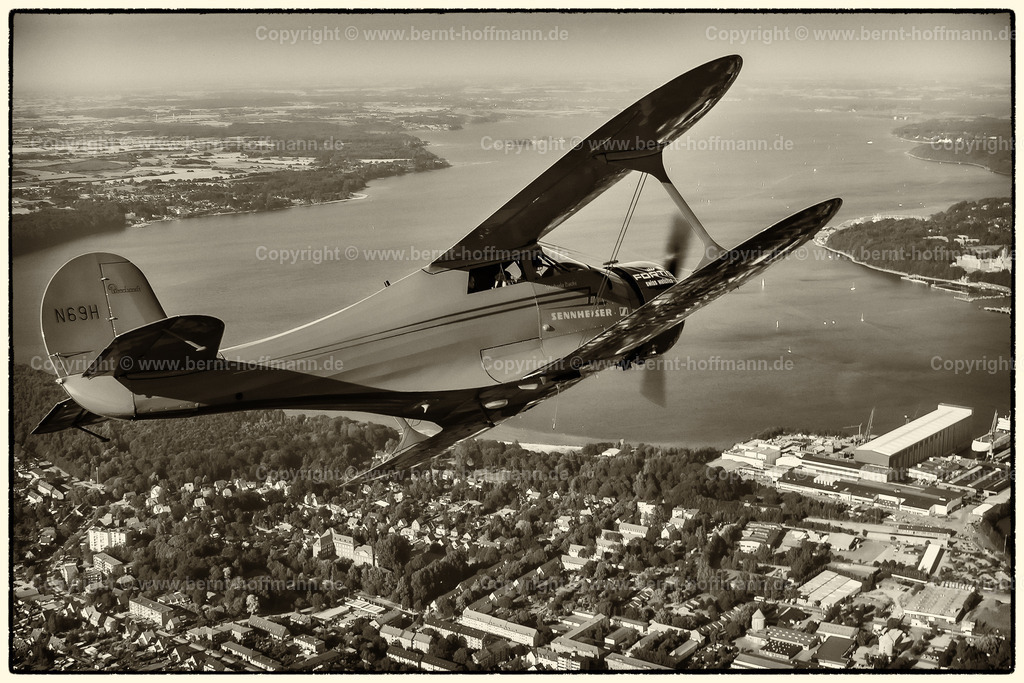 PLB_air2air_118_1827modnk1_75x50 | Luftbild. Historischer Doppeldecker Typ Beechcraft Staggerwing über Flensburg. Im Bildvordergrund - die Flensburger Nordstadt mit der Werft ( FSG ) ___ Bildanmutung = Duoton ockerfarben, farbreduziert. - Realisiert mit Pictrs.com