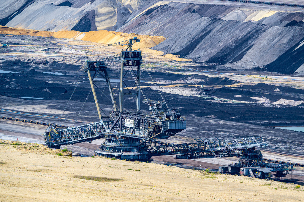 Industrie-Gigant im Schichtenwechsel: Absetzer im Tagebau | Das Foto zeigt einen gewaltigen Absetzer (Krupp/Siemens) inmitten einer kontrastreichen Tagebaulandschaft. Das massive Stahl-Großgerät steht im Zentrum auf dunklem, kohleartigen Untergrund und erstreckt seine Ausleger weit nach links und rechts. Der Hintergrund wird von monumentalen, terrassenförmig aufgeschütteten Erdwällen in verschiedenen Grau-, Braun- und Gelbtönen dominiert. Im hellen Vordergrund hebt sich eine sandige Ebene ab. Die Aufnahme fängt die beeindruckende Dimension moderner Industrietechnik und den massiven Eingriff in die Geologie ein. - Realisiert mit Pictrs.com