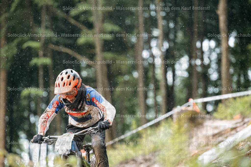 Mountainbike-Abenteuer im Wald | Ein Mountainbiker rast einen staubigen, schmalen Pfad in einem dichten Wald hinunter. Der Fahrer trägt einen orangefarbenen Helm und Schutzkleidung und fährt ein Mountainbike mit Federgabel. Der Hintergrund besteht aus dichtem Grün und Bäumen. Es regnet leicht. - Realisiert mit Pictrs.com