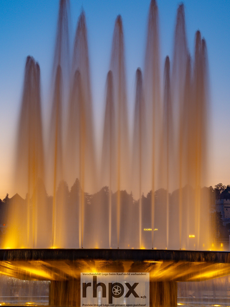 Wagenbach-Brunnen am KKL | Wasser. Licht. Eleganz.
Die illuminierten Wasserfontänen des Wagenbach-Brunnens steigen vor der modernen Kulisse des KKL in den Abendhimmel empor. Die goldene Beleuchtung kontrastiert sanft mit dem kühlen Blau der Dämmerung und schafft eine fast meditative Dynamik.
Wähle unter "Produktauswahl" dein Wunschformat: Vom klassischen Wandbild über Puzzle &amp; Tassen bis zum digitalen Download (z.B. als Handy-Hintergrund).
 - Realisiert mit Pictrs.com