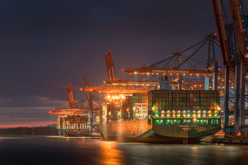 Nachts am Burchardkai Nr.1 | Das Bild zeigt eine nächtliche Szenerie im Hamburger Hafen am Burchardkai, wo mehrere riesige Containerschiffe angedockt sind. Die Schiffe erheben sich majestätisch in die Dunkelheit und werden von einer Flut von Lichtern beleuchtet, die vom Hafen und von den Schiffen selbst ausgehen. Das Bild fängt die Hektik und den Puls des Hafens in der Nacht ein und vermittelt einen Eindruck von der enormen Größe und dem Einfluss dieses wichtigen Handelszentrums.