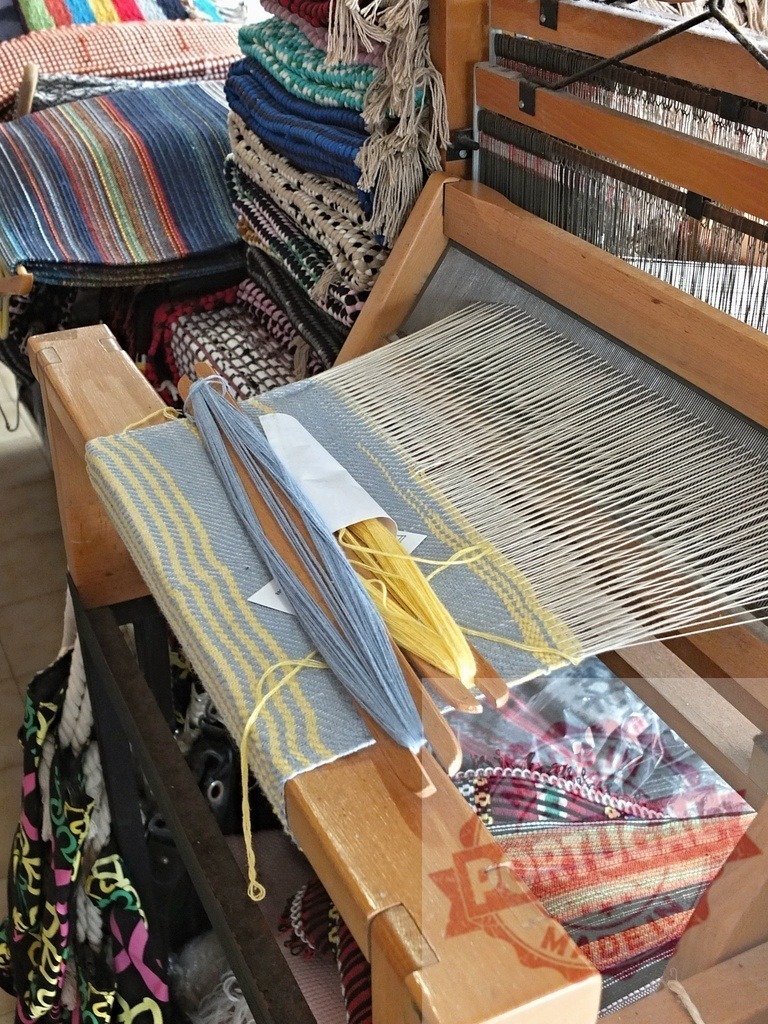 Traditional loom weaving colorful fabric and yarn shuttles | Weaving loom with yarn shuttles creating striped fabric next to stacked textiles - Realisiert mit Pictrs.com