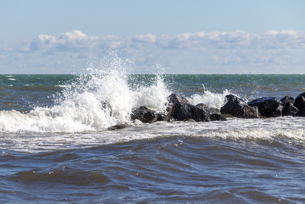 Wandbild: Brandung der stürmischen Ostsee | Dieses Wandbild im Querformat zeigt die stürmische Ostsee im Frühling. An der Steinmole bricht eine Welle. Am blauen Himmel sind einige Wolken zu sehen.  - Realisiert mit Pictrs.com
