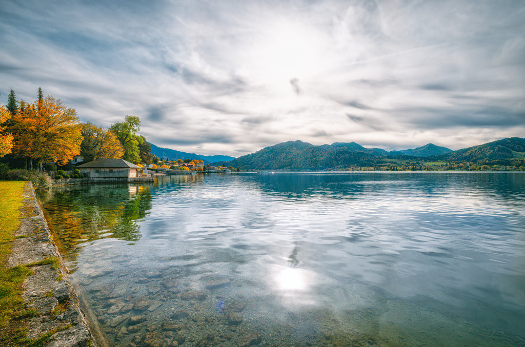 Blick über den Tegernsee und ein Bootshaus umrahmt von herbstlich gefärbten Bäumen | Das ausgewählte Bild zeigt einen malerischen Blick auf den Tegernsee, einen der bekanntesten und beliebtesten Voralpenseen in Oberbayern. Die Fotografie fängt die herbstliche Atmosphäre an den Ufern des Sees ein, während die Sonne durch ein wolkenverhangenes Himmelszelt bricht und sich auf der leicht welligen Wasseroberfläche spiegelt.Auf der linken Bildseite ist das im Webseiten-Titel beschriebene, charakteristische hölzerne Bootshaus zu sehen, welches von Bäumen in bunter Herbstfärbung umrahmt wird. Im Hintergrund erstreckt sich die Silhouette der bayerischen Voralpen (Mangfallgebirge), die das Tal des Sees umschließen. Der Tegernsee ist ein bekanntes Ausflugsziel südlich von München und zieht Besucher für Aktivitäten wie Wandern, Bootstouren und traditionelle bayerische Gasthauskultur an. - Realisiert mit Pictrs.com