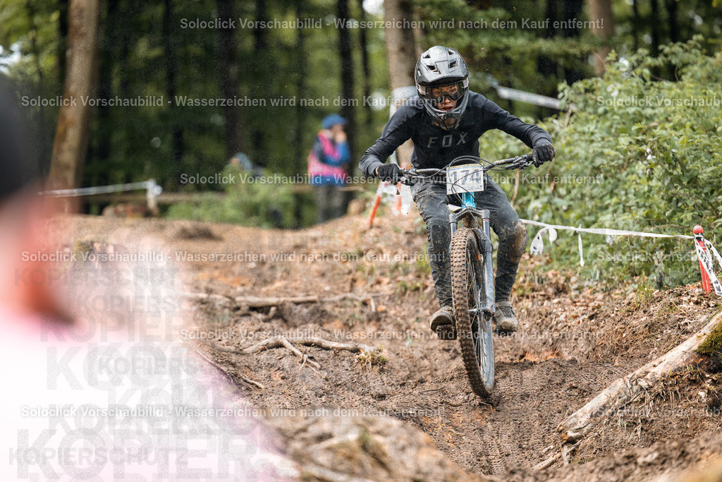 Mountainbiker im Wald | Das Foto zeigt einen Mountainbiker in voller Ausrüstung, der einen steilen, schlammigen Trail in einem dichten Wald hinunterfährt. Der Fahrer trägt einen schwarzen Helm mit einer roten Visier, eine dunkle Brille, eine schwarze Jacke mit dem Schriftzug 'FOX' und schwarze Handschuhe. Er trägt die Nummer 74 auf seinem Rücken. Der Trail ist mit Wurzeln und Steinen übersät und der Boden ist mit Schlamm bedeckt. Im Hintergrund sind weitere Bäume und ein Zuschauer zu sehen. Das Wetter scheint regnerisch zu sein. - Realisiert mit Pictrs.com