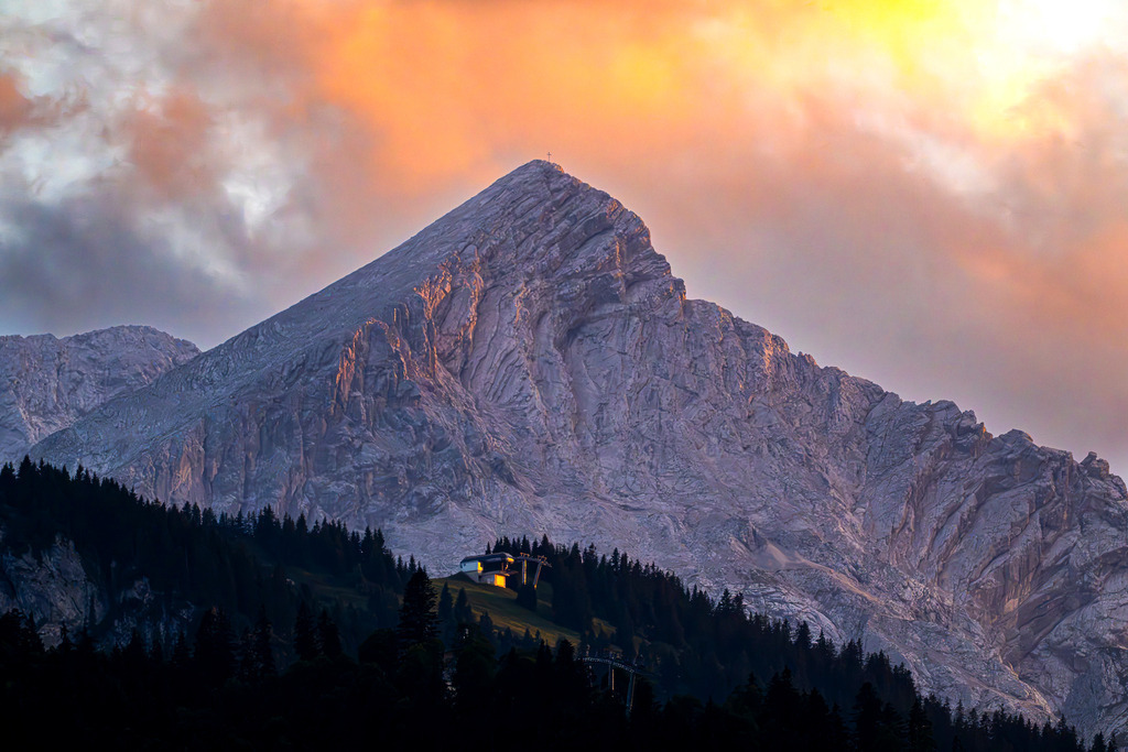 Magisches Alpenglühen: Der Alpspitz-Gipfel im Abendlicht | Ein erhabener Naturmoment über Garmisch-Partenkirchen: Die markante Pyramide der Alpspitze erstrahlt im spektakulären Licht der untergehenden Sonne. Goldene und roséfarbene Wolken hüllen den imposanten Felsgipfel in ein warmes, fast mystisches Glühen. Während der bewaldete Hang mit der Bergstation bereits sanft im Schatten liegt, thront das Gipfelkreuz stolz im letzten Sonnenlicht. Eine kraftvolle Aufnahme, die die wilde Ehrfurcht der Alpen, Erhabenheit und magische Bergmomente spürbar macht. - Realisiert mit Pictrs.com