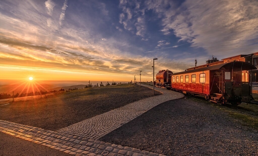 Schöner Sonnenaufgang auf dem Brocken-Harz--Sachsen Anhalt | Das Bild zeigt die Brockenbahn auf dem höchsten Berg im Harz, dem Brocken, während eines Sonnenaufgangs. Ort: Der Brockenbahnhof liegt auf einer Höhe von 1.125 Metern und ist der höchste Schmalspurbahnhof Deutschlands. Betrieb: Die Strecke wird von der Harzer Schmalspurbahnen GmbH (HSB) mit historischen Dampflokomotiven befahren. Erlebnis: Eine Fahrt mit der Bahn gilt als Höhepunkt eines Urlaubs im Harz und bietet ein einzigartiges Eisenbahnerlebnis. Wetterabhängigkeit: Bei extremen Wetterbedingungen wie Orkanböen wird der Zugverkehr zum Gipfel eingestellt.  - Realisiert mit Pictrs.com