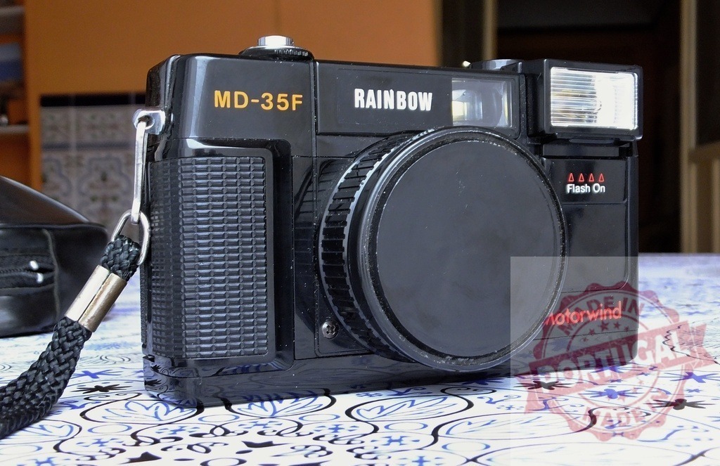 Rainbow MD-35F vintage 35mm film camera on table | Black retro compact film camera with flash and lens cap resting on a patterned tablecloth - Realisiert mit Pictrs.com