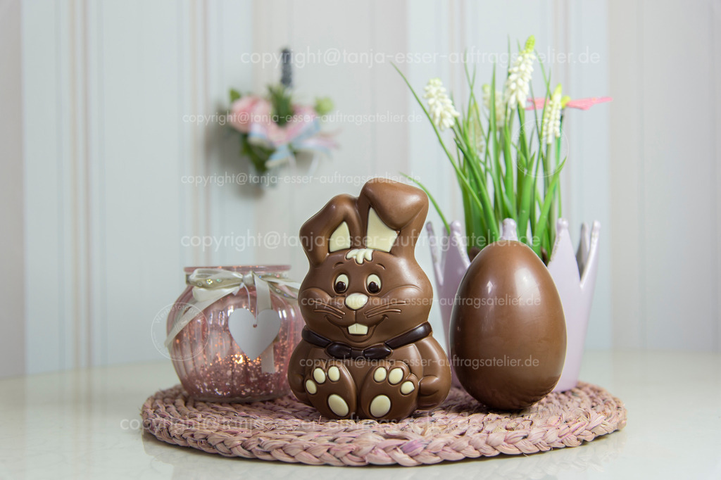 Schokoladen Hase und Ei 2 D | Ein Schokoladen Hase und ein Schokoladen Ei stehen auf einem Tisch, der mit Hyazinthen und Kerzen dekoriert ist. Verschwommene pinke Deko Elemente. 