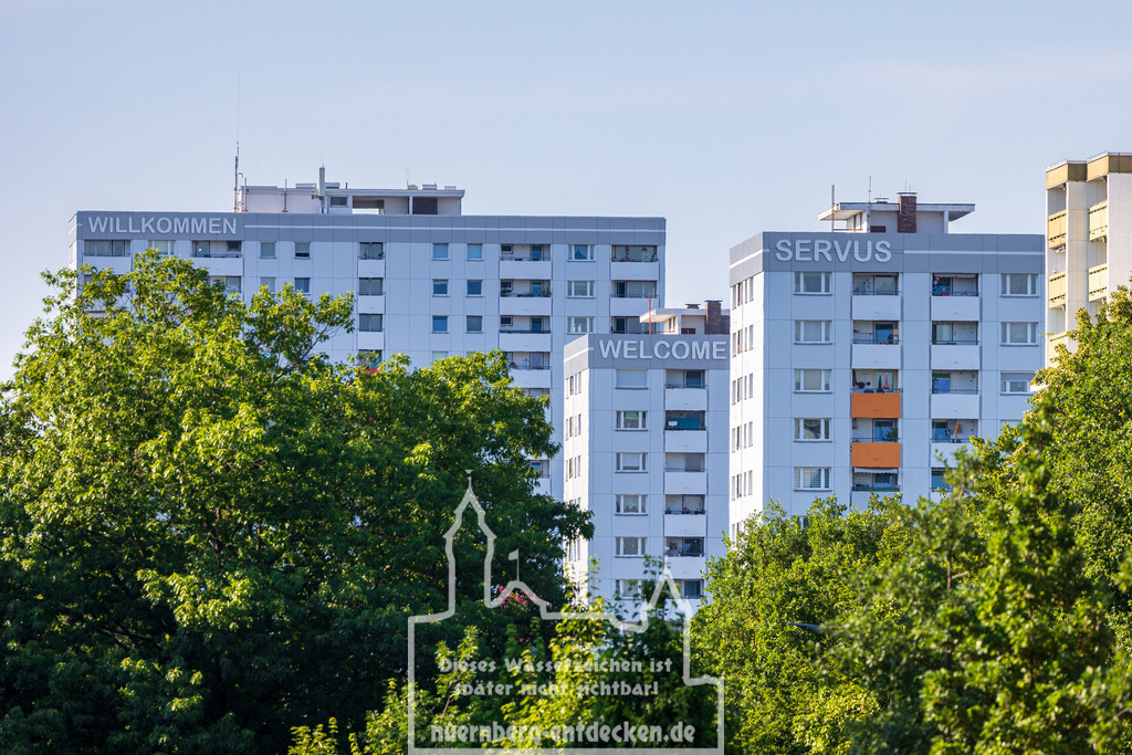 Monatsbild des Kalender 2026 für den Monat Juni | Mehrere Wohnhochhäuser mit grauer und weißer Fassade sind hinter einem dichten Grünbereich zu sehen. Auf den obersten Etagen der Gebäude sind Begrüßungsworte in großen Buchstaben angebracht: „WILLKOMMEN“, „WELCOME“ und „SERVUS“. Die Aufnahme zeigt die bekannte Willkommensansicht bei der Einfahrt nach Nürnberg von der Münchener Straße (B8) kommend. Die Gebäude gehören zum Stadtteil Langwasser. - Realisiert mit Pictrs.com