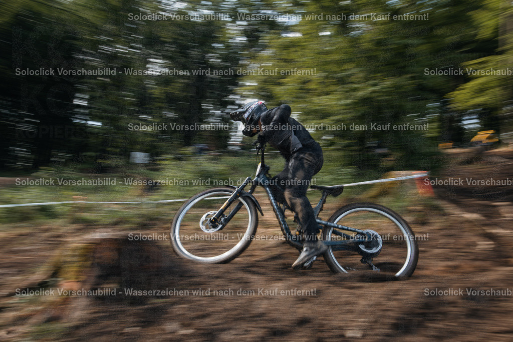 Dynamische Fahrt durch den Wald | Das Bild zeigt einen Mountainbiker in voller Montur, der mit hoher Geschwindigkeit durch einen verschwommenen Waldabschnitt fährt. Die Bewegung des Fahrers und des Fahrrads wird durch die lange Belichtungszeit deutlich hervorgehoben, was einen dynamischen Effekt erzeugt. Der Hintergrund besteht aus dichtem Grün und braunen Erdlagen. - Realisiert mit Pictrs.com