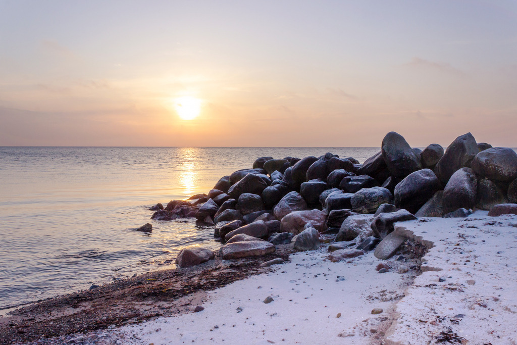 Wandbild: Sonnenaufgang am verschneiten Sandstrand | Dieses Wandbild im Querformat zeigt einen verschneiten Sandstrand im morgendlichen Licht der aufgehenden Sonne. Die Steine der Steinmole sind ebenfalls mit einer leichten Schneeschicht bedeckt. Die Sonne taucht den Himmel am Horizont in ein dezentes orange.  - Realisiert mit Pictrs.com
