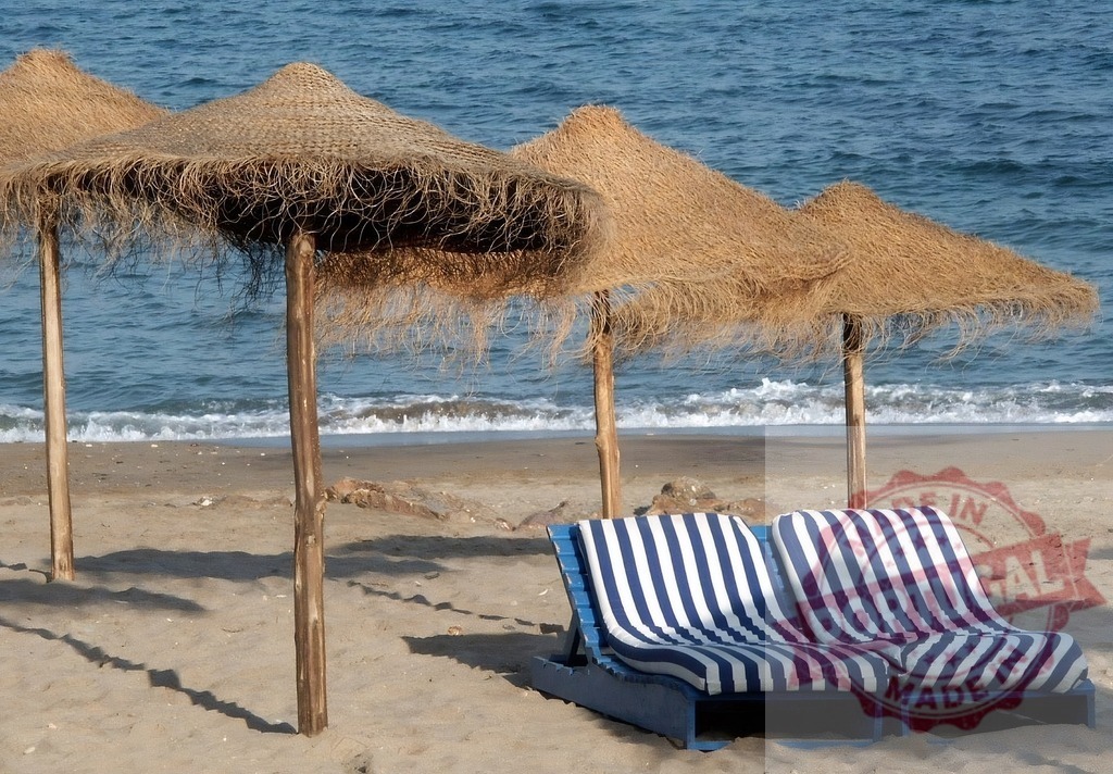 Straw umbrellas providing shade over beach chairs beside ocean. Espana - Cadiz | Straw beach umbrellas shading striped lounge chairs on hot sand by the sea - Realized with Pictrs.com